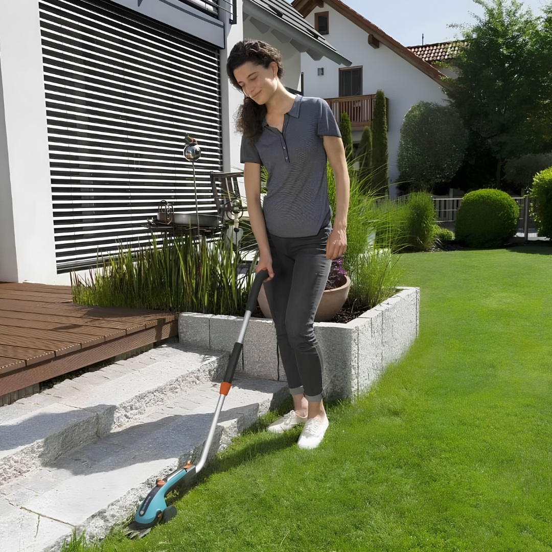 Akumulátorové nožnice na trávu gardena classiccut súprava 02 | záhradníctvo garden team Woman in a gray polo trims the edge of a stone walkway with a string trimmer beside a modern house in a sunny backyard