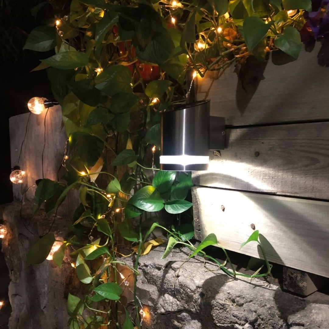 Nástenná led lampa garden na stenu 02 | záhradníctvo garden team Strieborné valcové vonkajšie svetlo svietiace na drevený obklad obklopený popínavými listami a žiarovkami